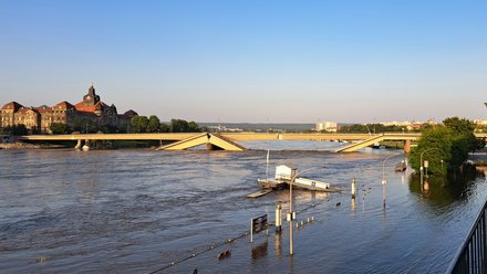 Eine Fotografie zeigt eine teilweise eingestürzte Brücke über einen stark angeschwollenen Fluss, Brückenteile sind abgesackt und ragen schräg ins Wasser, überschwemmte Uferbereiche und versunkene Infrastruktur verdeutlichen die Ausnahmesituation, im Hintergrund ist ein markantes historisches Gebäude sichtbar, wodurch das Bild die Verletzlichkeit technischer Bauwerke zeigt und für die Bedeutung von Instandhaltung, Resilienz und verantwortungsvollem Umgang mit bestehender Infrastruktur steht.