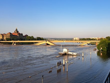 Eine Fotografie zeigt eine teilweise eingestürzte Brücke über einen stark angeschwollenen Fluss, Brückenteile sind abgesackt und ragen schräg ins Wasser, überschwemmte Uferbereiche und versunkene Infrastruktur verdeutlichen die Ausnahmesituation, im Hintergrund ist ein markantes historisches Gebäude sichtbar, wodurch das Bild die Verletzlichkeit technischer Bauwerke zeigt und für die Bedeutung von Instandhaltung, Resilienz und verantwortungsvollem Umgang mit bestehender Infrastruktur steht.