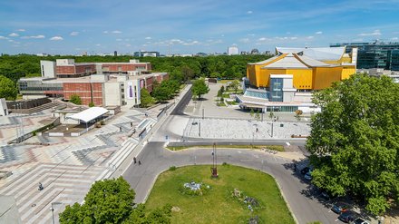 "Luftaufnahme eines großen öffentlichen Platzes mit gepflasterten Bereichen und Grünanlagen. Links ein rotbraunes modernes Gebäude mit Treppen und Bühnenbereich. Rechts ein markantes gelbes Gebäude mit weißem Dach und orangefarbenem Kunstwerk. Mehrere Personen bewegen sich auf dem Platz. Im Hintergrund grüne Parks, Bäume und Hochhäuser einer Stadtsilhouette unter blauem Himmel."