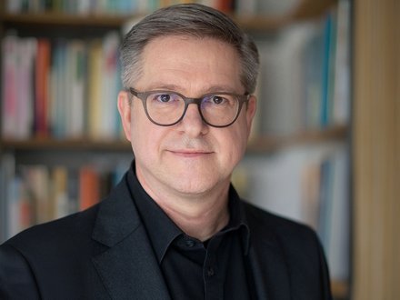 Porträtfoto einer mittelalten Person mit kurz grau melierten Haaren und runder Brille, im dunklen Sakko und schwarzem Hemd; neutraler Blick, Kopf-und-Schultern-Aufnahme vor unscharfem Bücherregal im Hintergrund.