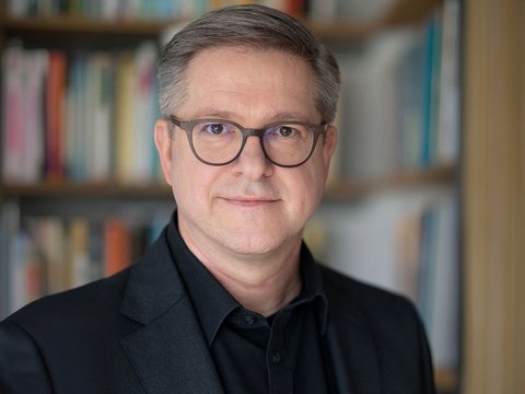 Porträtfoto einer mittelalten Person mit kurz grau melierten Haaren und runder Brille, im dunklen Sakko und schwarzem Hemd; neutraler Blick, Kopf-und-Schultern-Aufnahme vor unscharfem Bücherregal im Hintergrund.