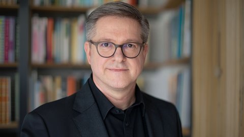 Porträtfoto einer mittelalten Person mit kurz grau melierten Haaren und runder Brille, im dunklen Sakko und schwarzem Hemd; neutraler Blick, Kopf-und-Schultern-Aufnahme vor unscharfem Bücherregal im Hintergrund.