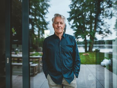 Mann mit dunklem Hemd und beiger Hose steht vor einer Glasfront mit Blick auf Garten und Bäume.