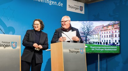 Zwei Personen stehen an einem Rednerpult mit dem Schriftzug 'Freistaat Thüringen' vor einer blauen Wand mit dem Schriftzug 'thueringen.de'. Neben ihnen ist ein Bildschirm mit einem Bild eines Gebäudes und dem Text 'Regierungsmedienkonferenz der Thüringer Staatskanzlei'.