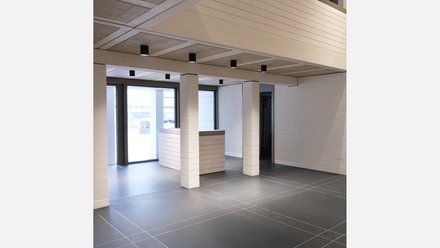 Foyer mit niedriger Empfangstheke, hellen Ziegelpfeilern, großen Fenstern und Rasterdecke mit schwarzen Downlights; dunkler Boden.