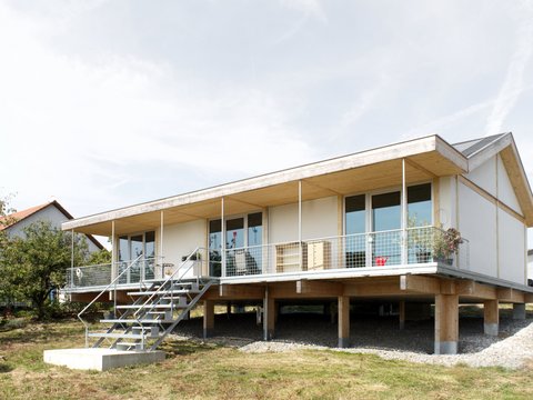 Außenansicht: eingeschossiges Haus auf Pfeilern mit Veranda und Metalltreppe; Putz‑ und Holzelemente, Satteldach.