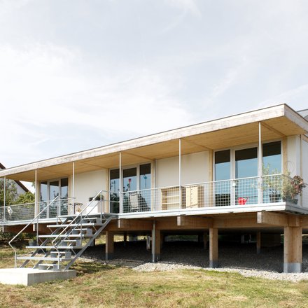 Außenansicht: eingeschossiges Haus auf Pfeilern mit Veranda und Metalltreppe; Putz‑ und Holzelemente, Satteldach.