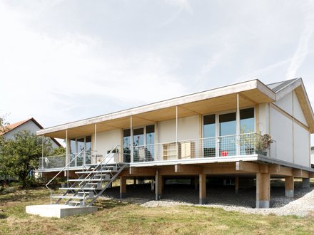 Außenansicht: eingeschossiges Haus auf Pfeilern mit Veranda und Metalltreppe; Putz‑ und Holzelemente, Satteldach.