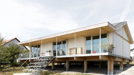 Außenansicht: eingeschossiges Haus auf Pfeilern mit Veranda und Metalltreppe; Putz‑ und Holzelemente, Satteldach.
