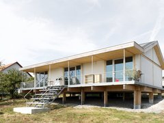 Außenansicht: eingeschossiges Haus auf Pfeilern mit Veranda und Metalltreppe; Putz‑ und Holzelemente, Satteldach.