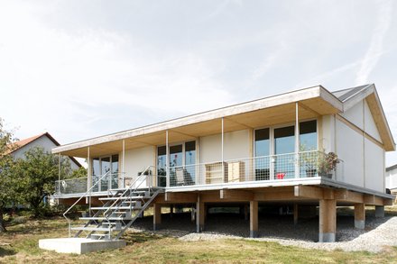 Außenansicht: eingeschossiges Haus auf Pfeilern mit Veranda und Metalltreppe; Putz‑ und Holzelemente, Satteldach.