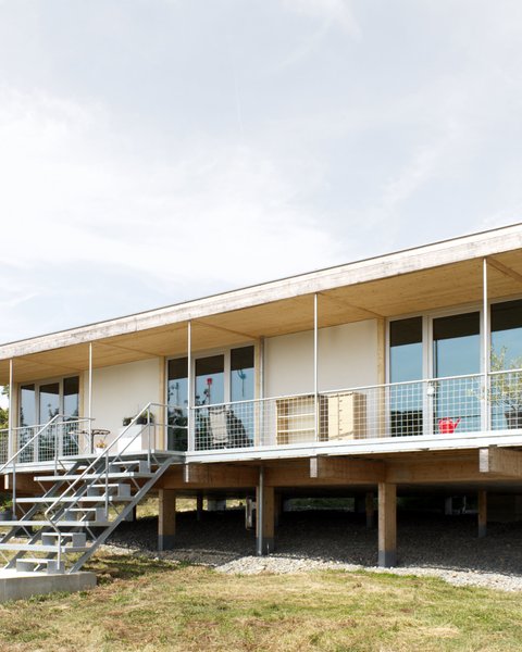 Außenansicht: eingeschossiges Haus auf Pfeilern mit Veranda und Metalltreppe; Putz‑ und Holzelemente, Satteldach.