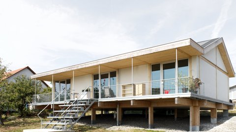 Außenansicht: eingeschossiges Haus auf Pfeilern mit Veranda und Metalltreppe; Putz‑ und Holzelemente, Satteldach.