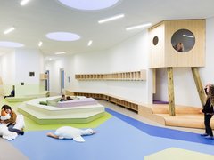 Kindertagesstätte mit modernem, hellem Innenraum, Spielflächen in verschiedenen Ebenen und einem hölzernen Spielhaus auf Stelzen mit rundem Fenster