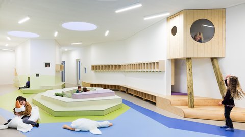 Kindertagesstätte mit modernem, hellem Innenraum, Spielflächen in verschiedenen Ebenen und einem hölzernen Spielhaus auf Stelzen mit rundem Fenster