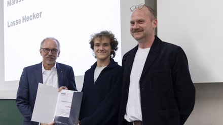 Drei Personen bei einer Auszeichnung; die mittlere Person zeigt eine Urkunde mit der Aufschrift „1. Preis“.