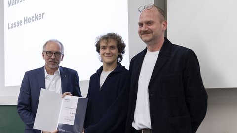 Drei Personen bei einer Auszeichnung; die mittlere Person zeigt eine Urkunde mit der Aufschrift „1. Preis“.