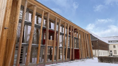 Modernes Holzgebäude mit charakteristischer Säulenvorhalle aus massiven Holzpfosten. Die Fassade besteht aus hellen Holzplanken mit großflächigen Fenster- und Glastürbereichen. Eine dunkelrote Tür ist sichtbar. Der Boden ist mit Schnee bedeckt, davor stehen Absperrkegel. Im Hintergrund mehrgeschossige Gebäude aus Backstein und Holz. Blauer Himmel mit Wolken. Das Gebäude wirkt wie ein modernes öffentliches oder kulturelles Gebäude.