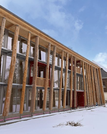 Modernes Holzgebäude mit charakteristischer Säulenvorhalle aus massiven Holzpfosten. Die Fassade besteht aus hellen Holzplanken mit großflächigen Fenster- und Glastürbereichen. Eine dunkelrote Tür ist sichtbar. Der Boden ist mit Schnee bedeckt, davor stehen Absperrkegel. Im Hintergrund mehrgeschossige Gebäude aus Backstein und Holz. Blauer Himmel mit Wolken. Das Gebäude wirkt wie ein modernes öffentliches oder kulturelles Gebäude.