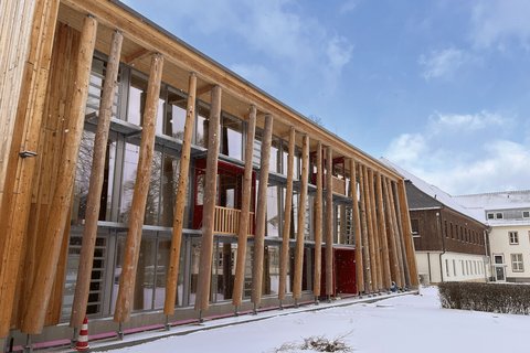 Modernes Holzgebäude mit charakteristischer Säulenvorhalle aus massiven Holzpfosten. Die Fassade besteht aus hellen Holzplanken mit großflächigen Fenster- und Glastürbereichen. Eine dunkelrote Tür ist sichtbar. Der Boden ist mit Schnee bedeckt, davor stehen Absperrkegel. Im Hintergrund mehrgeschossige Gebäude aus Backstein und Holz. Blauer Himmel mit Wolken. Das Gebäude wirkt wie ein modernes öffentliches oder kulturelles Gebäude.