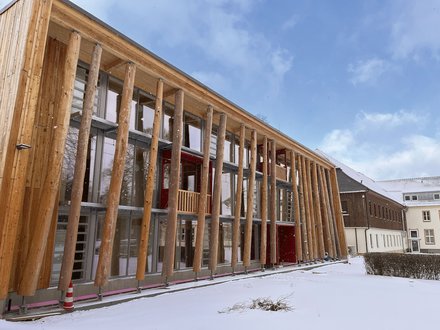 Modernes Holzgebäude mit charakteristischer Säulenvorhalle aus massiven Holzpfosten. Die Fassade besteht aus hellen Holzplanken mit großflächigen Fenster- und Glastürbereichen. Eine dunkelrote Tür ist sichtbar. Der Boden ist mit Schnee bedeckt, davor stehen Absperrkegel. Im Hintergrund mehrgeschossige Gebäude aus Backstein und Holz. Blauer Himmel mit Wolken. Das Gebäude wirkt wie ein modernes öffentliches oder kulturelles Gebäude.