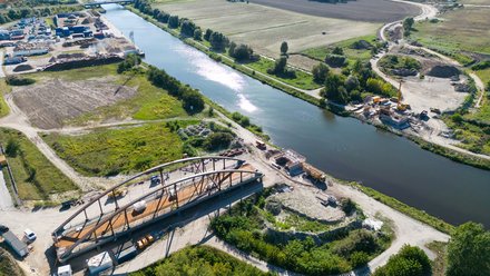 Weite Luftaufnahme eines Kanals mit Baustelle: neue Stahlbogenbrücke entsteht über dem Wasser; Kräne, Widerlager und Baustraßen sichtbar, umgeben von Feldern und Gewerbeflächen.