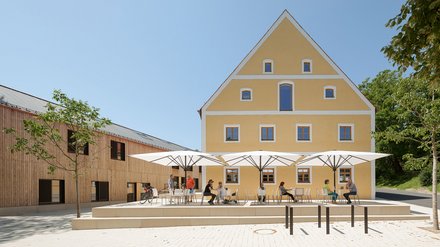 "Gelbes Hauptgebäude mit Satteldach und blauen Fensterrahmen, flankiert von Holzgebäuden. Im befestigten Innenhof sitzen mehrere Personen unter weißen Sonnenschirmen an Tischen. Grüne Bäume rahmen das Gelände ein. Klarer blauer Himmel."