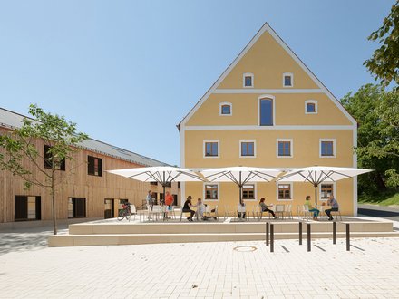"Gelbes Hauptgebäude mit Satteldach und blauen Fensterrahmen, flankiert von Holzgebäuden. Im befestigten Innenhof sitzen mehrere Personen unter weißen Sonnenschirmen an Tischen. Grüne Bäume rahmen das Gelände ein. Klarer blauer Himmel."
