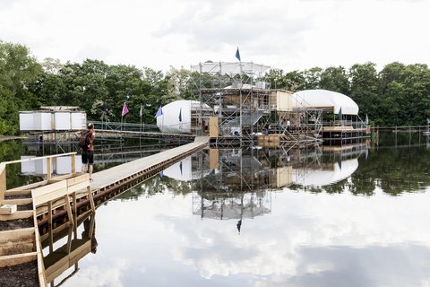 Konstruktion aus Gerüst, Holz und weißen Kuppeln auf einem See mit Steg und Person mit Rucksack