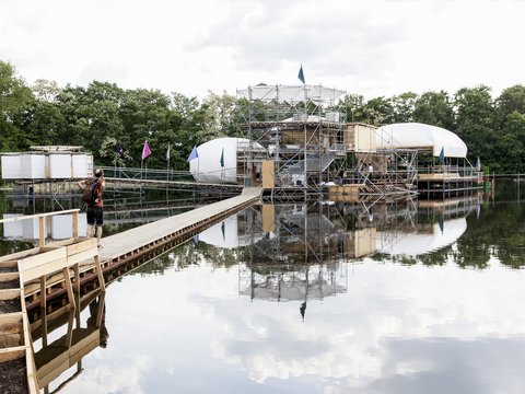 Konstruktion aus Gerüst, Holz und weißen Kuppeln auf einem See mit Steg und Person mit Rucksack
