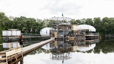 Konstruktion aus Gerüst, Holz und weißen Kuppeln auf einem See mit Steg und Person mit Rucksack