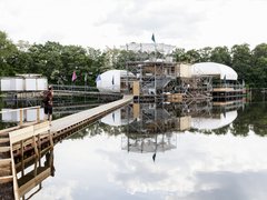 Konstruktion aus Gerüst, Holz und weißen Kuppeln auf einem See mit Steg und Person mit Rucksack