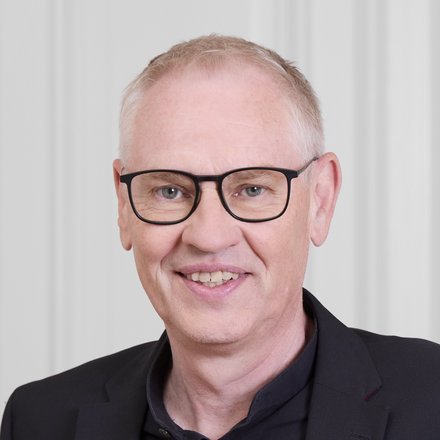 Halbnahes Farbfoto einer lächelnden erwachsenen Person mit Kurzhaar und Brille, in schwarzem Hemd und Sakko, vor heller, klassischer Türwand; Blick in die Kamera.