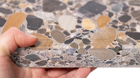 Hand hält eine Terrazzo-Platte; bunte Kiesel im grauen Bindemittel, Kante im Querschnitt sichtbar.