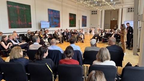 "Großer Veranstaltungssaal mit heller Architektur und modernem Leuchtenkunstwerk an der Decke. Etwa 50 Personen sitzen in einem Stuhlkreis auf Holzboden. An den Wänden hängen vier abstrakte Kunstwerke mit grünem und blauem Hintergrund und roten geometrischen Formen. Im Hintergrund stehen zwei Personen neben einer Präsentationsleinwand. Die Veranstaltung wirkt wie ein Diskussionsforum oder Workshop in professionellem Rahmen."