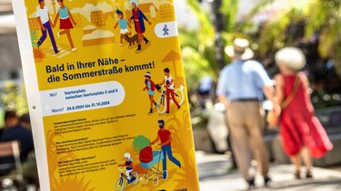 Banner mit Aufschrift: Bald in Ihrer Nähe - die Sommerstraße kommt! Mit Fußgängern im Hintergrund. 