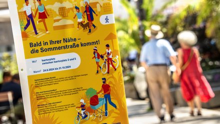 Banner mit Aufschrift: Bald in Ihrer Nähe - die Sommerstraße kommt! Mit Fußgängern im Hintergrund. 