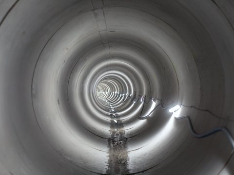 "Blick durch einen großen zylindrischen Betonabwassertunnel. Konzentrische Ringe verlaufen perspektivisch zum Lichtpunkt in der Ferne. Eine dünne Wasserschicht fließt durch den Tunnelboden. Seitlich tritt Licht von rechts ein. Teil des Kopenhagener Regenwassermanagementsystems."
