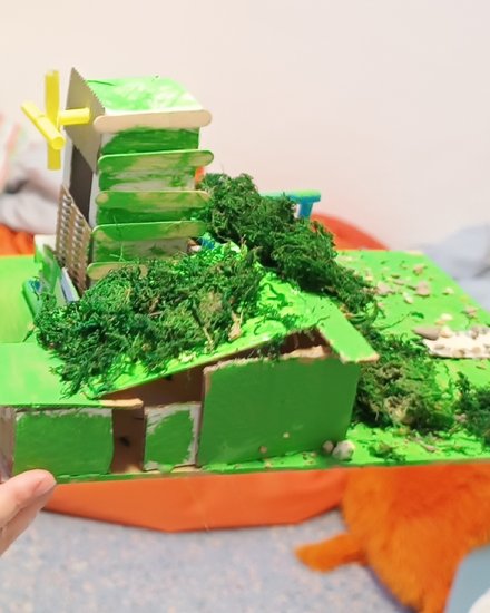 "Detailliertes 3D-Modell einer grünen Stadtlandschaft aus Karton und Moos. Eine Person hält das Modell in den Händen. Das Modell zeigt grüne Gebäude, ein gelbes Kreuz auf einem Dach, künstliches grünes Moos als Vegetation, beige und grüne Strukturen. Im Hintergrund bunte Papierfahnen in Regenbogenfarben. Das Modell zeigt nachhaltiges, grünes Stadtplanen."