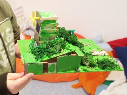 "Detailliertes 3D-Modell einer grünen Stadtlandschaft aus Karton und Moos. Eine Person hält das Modell in den Händen. Das Modell zeigt grüne Gebäude, ein gelbes Kreuz auf einem Dach, künstliches grünes Moos als Vegetation, beige und grüne Strukturen. Im Hintergrund bunte Papierfahnen in Regenbogenfarben. Das Modell zeigt nachhaltiges, grünes Stadtplanen."