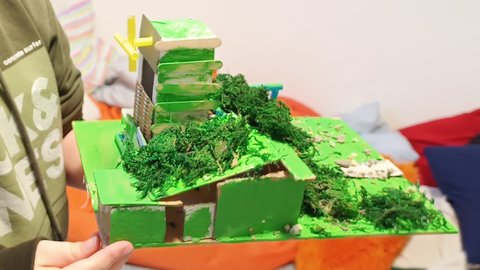 "Detailliertes 3D-Modell einer grünen Stadtlandschaft aus Karton und Moos. Eine Person hält das Modell in den Händen. Das Modell zeigt grüne Gebäude, ein gelbes Kreuz auf einem Dach, künstliches grünes Moos als Vegetation, beige und grüne Strukturen. Im Hintergrund bunte Papierfahnen in Regenbogenfarben. Das Modell zeigt nachhaltiges, grünes Stadtplanen."