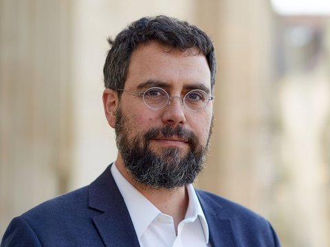"Porträtfoto eines Mannes mit dunklem, vollem Bart und dunklen Haaren. Er trägt eine dunkelblau-graue Anzugjacke über einem weißen Hemd und eine silberne Brille mit runder Fassung. Der Blick ist direkt zur Kamera gerichtet, der unscharfe Hintergrund deutet auf eine urbane Umgebung hin."