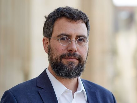"Porträtfoto eines Mannes mit dunklem, vollem Bart und dunklen Haaren. Er trägt eine dunkelblau-graue Anzugjacke über einem weißen Hemd und eine silberne Brille mit runder Fassung. Der Blick ist direkt zur Kamera gerichtet, der unscharfe Hintergrund deutet auf eine urbane Umgebung hin."
