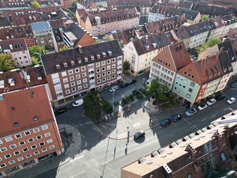 Luftaufnahme eines Stadtplatzes von oben. Mehrgeschossige historische Gebäude mit roten und braunen Ziegeldächern umrahmen den Platz. Graue Asphaltfläche mit geparkten Autos. Kleine Grünflächen und vereinzelte Bäume. Mehrere Fußgänger auf der Fläche.