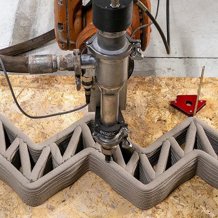 Roboterbasierter 3D-Druckkopf extrudiert Lehm in einer Zickzack-Struktur und erstellt eine Wand mit inneren Verstrebungen auf einer Werkbank.