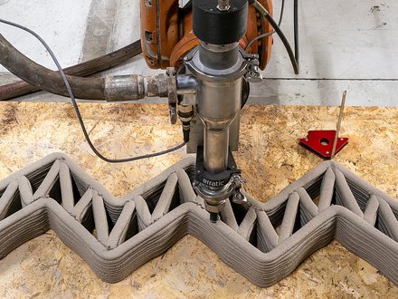 Roboterbasierter 3D-Druckkopf extrudiert Lehm in einer Zickzack-Struktur und erstellt eine Wand mit inneren Verstrebungen auf einer Werkbank.