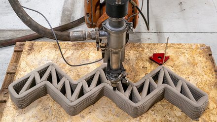 Roboterbasierter 3D-Druckkopf extrudiert Lehm in einer Zickzack-Struktur und erstellt eine Wand mit inneren Verstrebungen auf einer Werkbank.