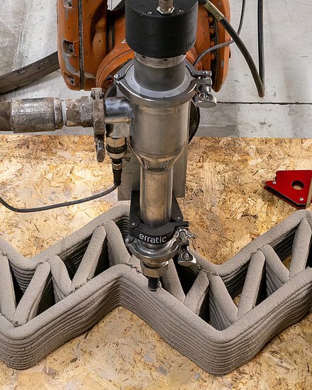 Roboterbasierter 3D-Druckkopf extrudiert Lehm in einer Zickzack-Struktur und erstellt eine Wand mit inneren Verstrebungen auf einer Werkbank.