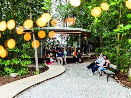 Belebter Garten um einen runden Pavillon; Menschen sitzen auf Bänken und am Steg, warme Lichtpunkte zwischen den Bäumen.