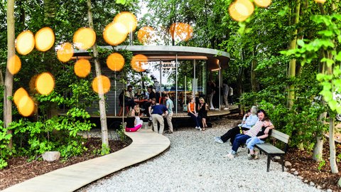 Belebter Garten um einen runden Pavillon; Menschen sitzen auf Bänken und am Steg, warme Lichtpunkte zwischen den Bäumen.